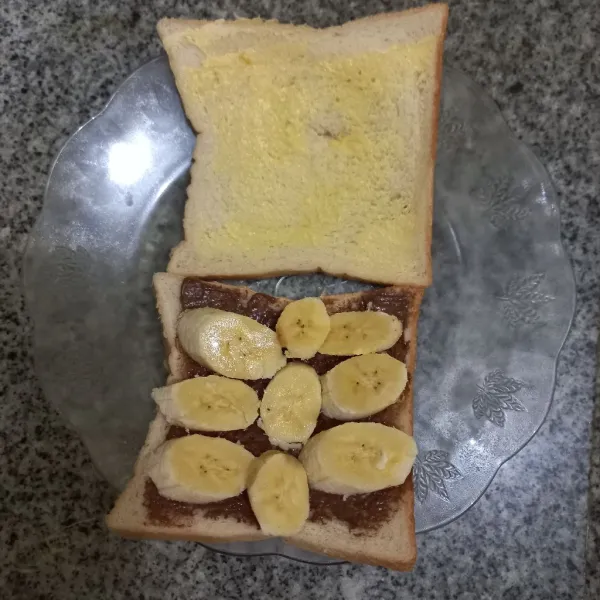 Tata potongan pisang di atas roti yang telah diolesi selai cokelat.