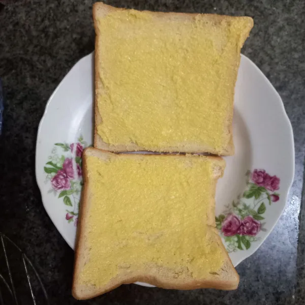 Olesi salah satu sisi kedua roti tawar dengan margarin.
