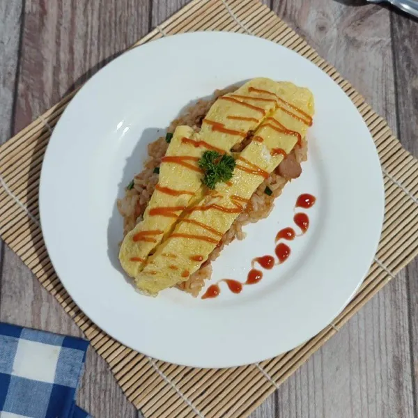 Tata nasi goreng memanjang di atas piring. Tambahkan omelette telur di atasnya. Belah bagian tengah telur (jika telur ½ matang dalamnya, begitu dibelah langsung menyelimuti nasi goreng). Sajikan selagi hangat.