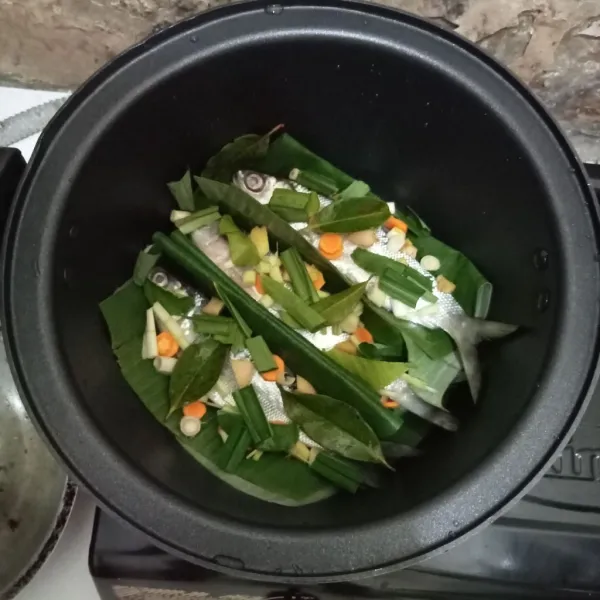 Tata ikan di presto. Alasi dengan daun pisang. Beri irisan kunyit, jahe, daun pandan, daun salam dan daun jeruk.