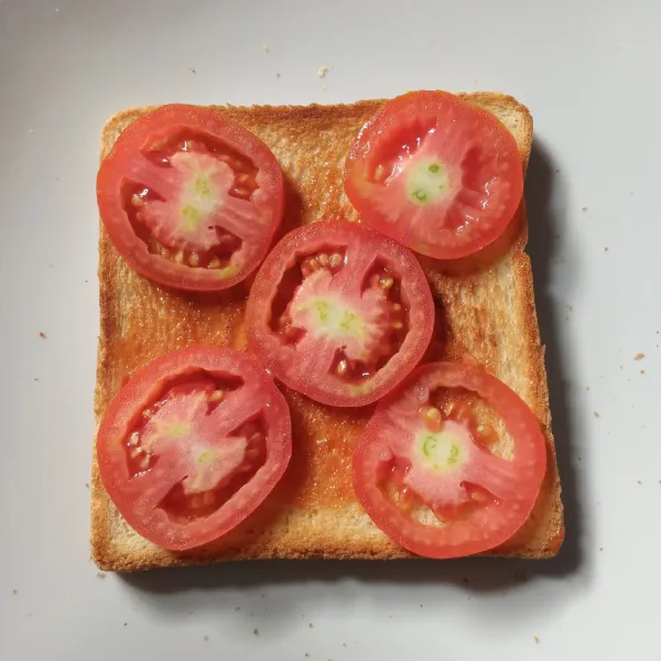 Ambil selembar roti panggang, oles dengan setengah sendok teh saus tomat. Tata potongan tomat.