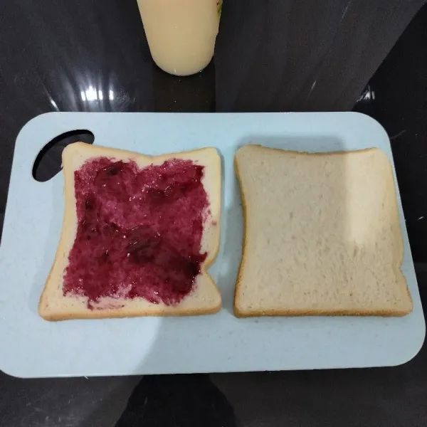 Oles rata salah satu sisi roti tawar dengan selai blueberry.