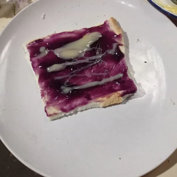 Ambil selembar roti tawar, olesi dengan selai blueberry dan kental manis.