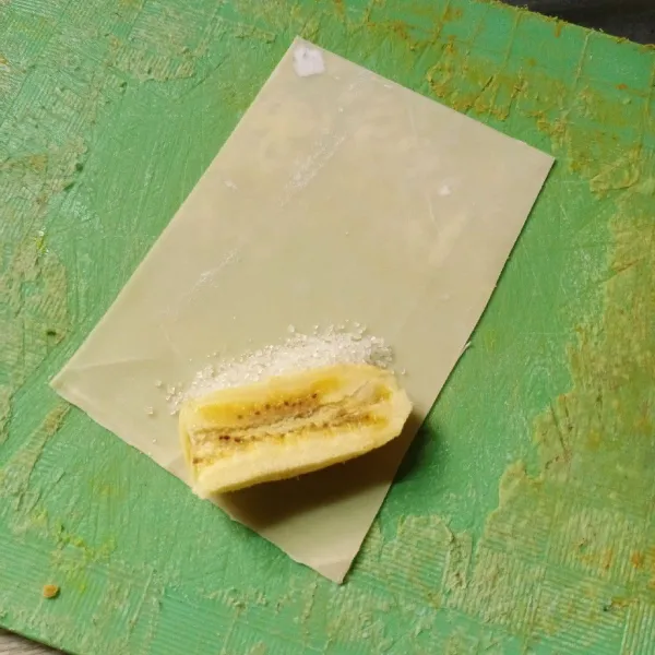 Ambil 1 lembar kulit pangsit, tata pisang dan sedikit gula pasir dibagian sudutnya. Kemudian lipat ujung kiri dan kanan lalu gulung. Rekatkan ujungnya dengan larutan terigu.