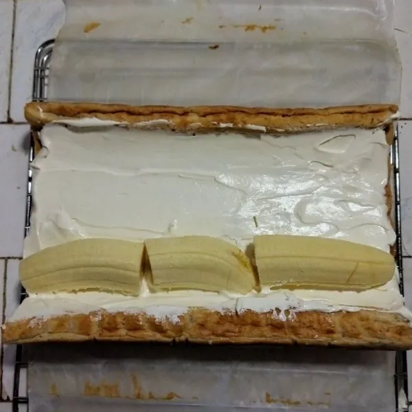Buka gulungan kue, lepaskan baking paper, oles filling dan tata pisang ambon. Dengan bantuan baking paper gulung kembali hingga padat dan rapi.