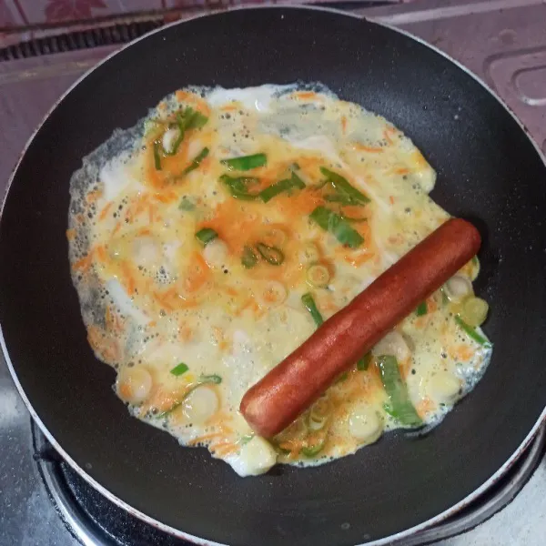 Panaskan teflon, olesi dengan sedikit minyak goreng, tuang adonan telur secukupnya, ratakan kemudian letakkan sosis di atasnya selagi telur belum matang.