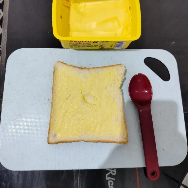 Oles salah satu sisi roti dengan margarin.