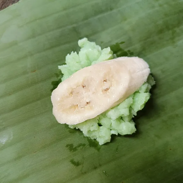 Letakkan dua sendok makan adonan dan sepotong pisang di atas daun pisang, lalu rapikan hingga pisang tertutup. adonan.