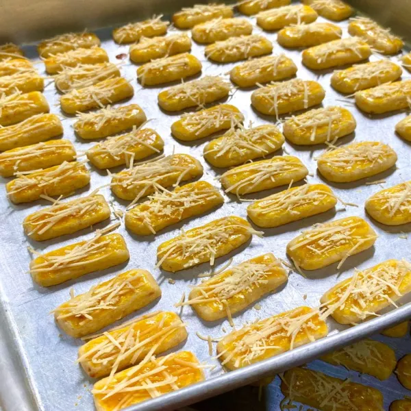 Siapkan loyang yang telah diberi margarin. Letakkan kastengel di atas loyang, tambahkan olesan kuning telur dan beri topping keju cheddar parut.
