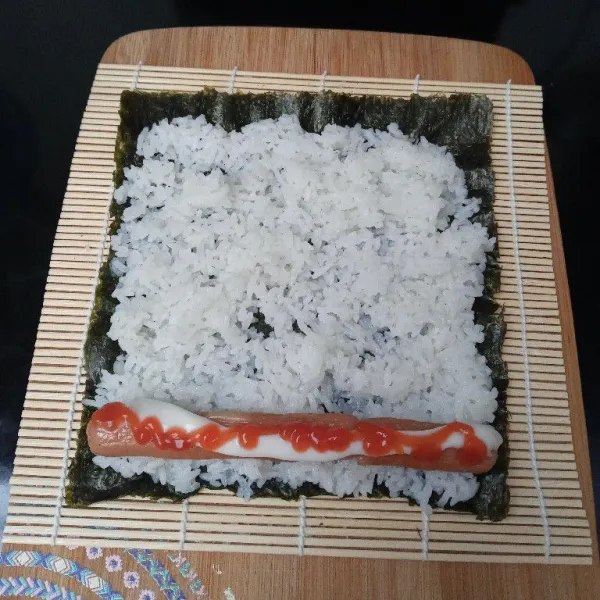 Taruh nasi di atas nori, ratakan, taruh sosis di salah satu ujungnya kemudian kasih mayonaise dan saus tomat