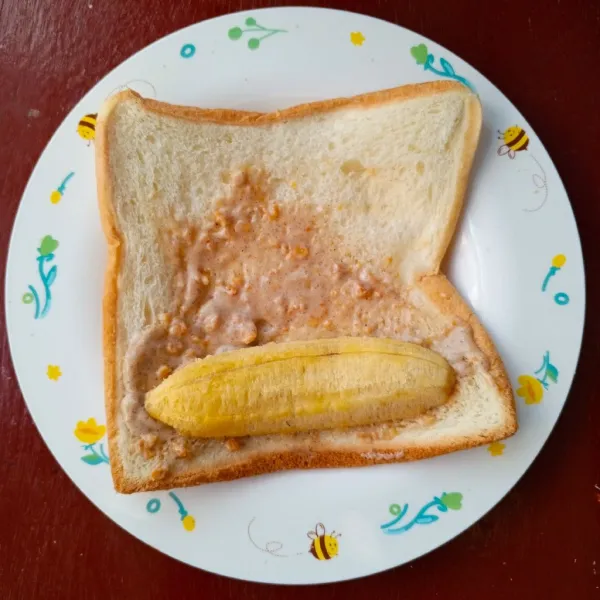 Oles roti dengan selai tiramisu crunchy, beri potongan pisang di atasnya.
