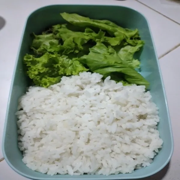 Tata nasi putih dan potongan daun selada dalam kotak bekal.