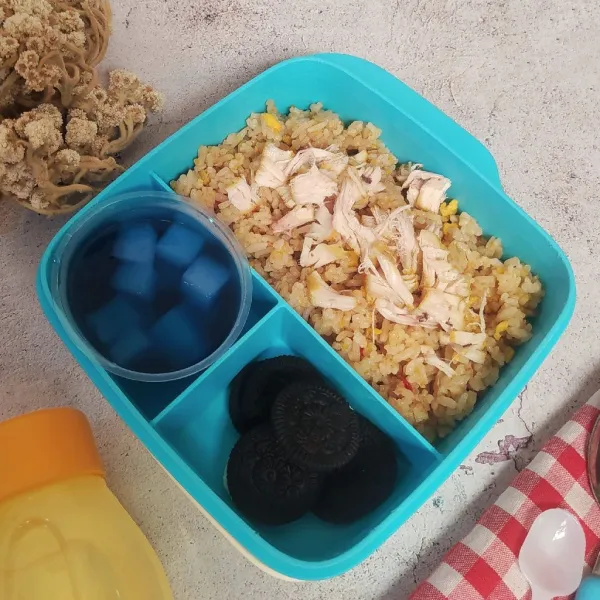 Tata nasi goreng di kotak bekal, beri taburan ayam suwir. Tambahkan puding, cookies dan air putih
