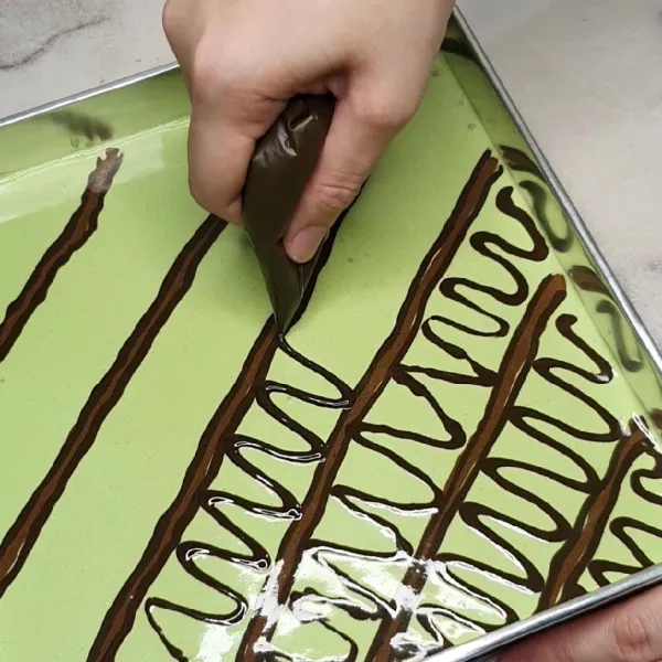 Tuang adonan pandan ke dalam loyang. Buat motif batik dengan adonan cokelat dan hitam.