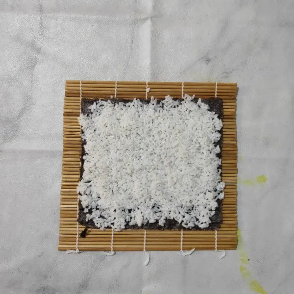 Siapkan sushi matt, letakkan nori di atas matt, kemudian tambahkan campuran nasi secara merata.