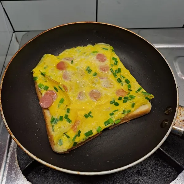 Jika bagian bawah sudah berkulit, balik telur dengan posisi roti di bawah. Lipat pinggiran telur agar rapi. Kecilkan api kompor.