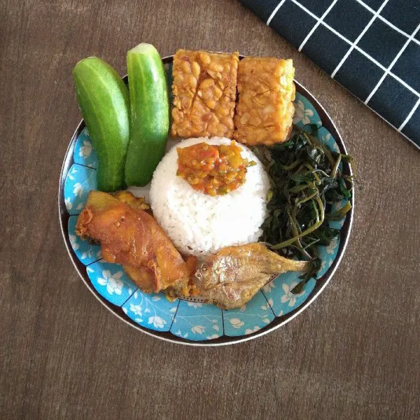 Tata nasi putih, ayam goreng, sayur rebus, timun, ikan asin dan sambal di piring, siap disajikan.