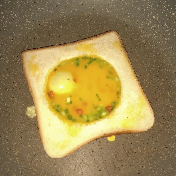 Panaskan teflon, dengan api kecil. Panggang roti, berikan margarin tepat dibaguan tengan roti yang bolong. Tuang setengah bagian telur. Tunggu sebentar.