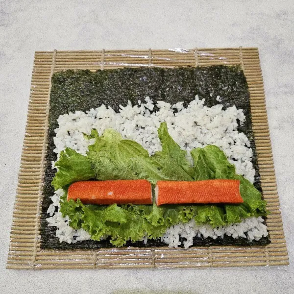 Tata nori di atas sushi mat. Beri nasi secukupnya, lalu ratakan. Setelah itu, tata selada dan crab stick di atas nasi