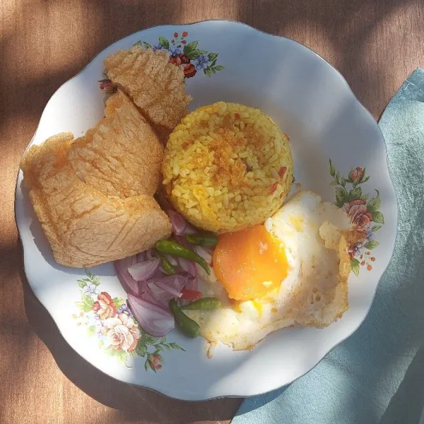 Tata nasi goreng kunyit kencur, telur ceplok, acar bawang merah dan kerupuk ketumbar di atas piring. Sajikan ketika masih hangat.