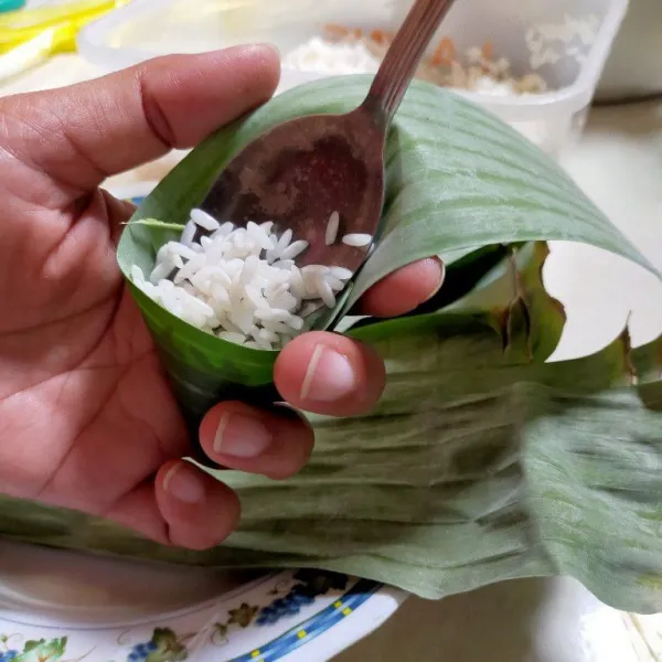 Potong daun memanjang dengan lebar 5 cm, bentuk segitiga kemudian isi dengan beras ketan.