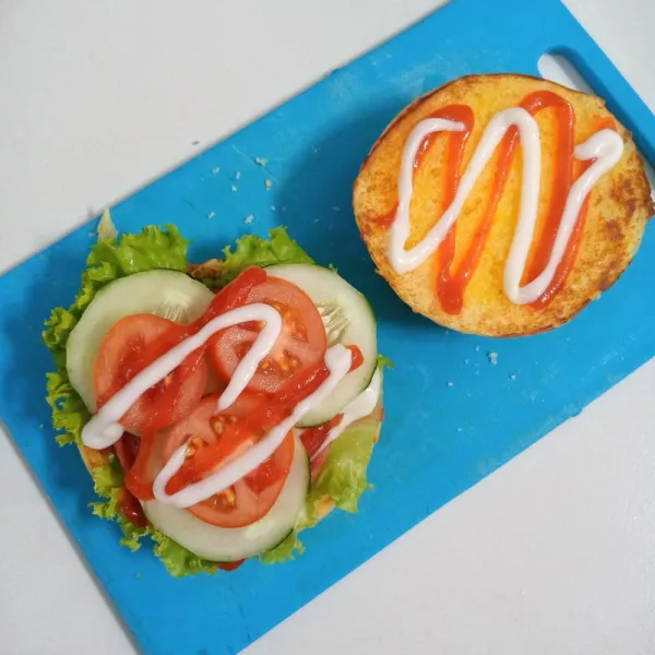 Tambahkan daun sla, smoked beef, timun, tomat, saos sambal dan mayonaise di atasnya kemudian tutup dengan roti. Burger siap disajikan.
