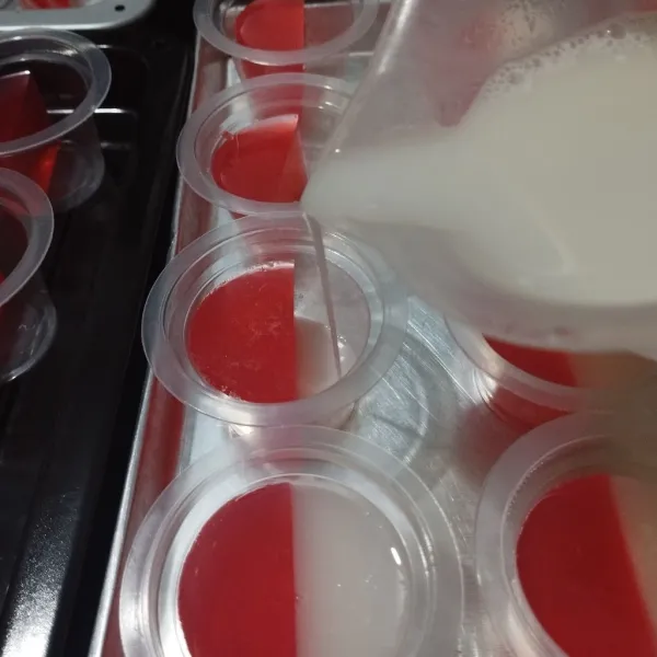 Tuang puding putih dalam cup yang sudah berisi separuh bagian puding merah, lakukan sampai selesai.