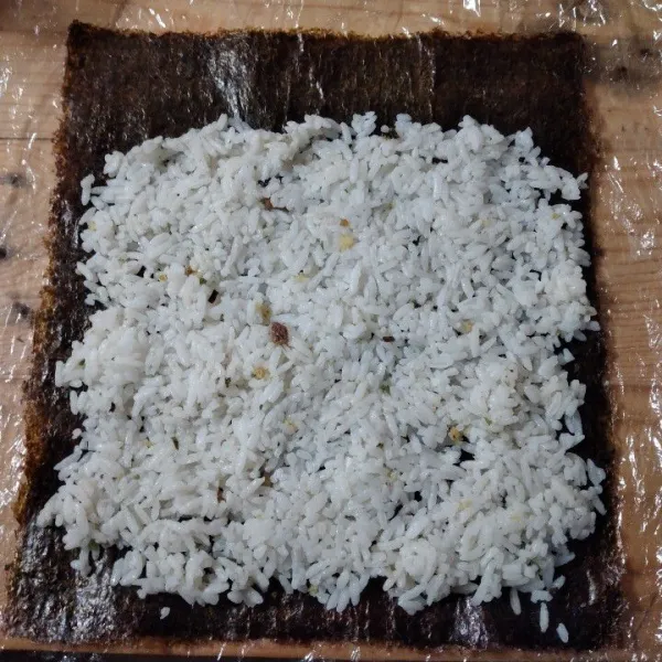 Alasi meja kerja dengan plastik, tata lembaran nori di atas plastik lalu ratakan nasi di atasnya.
