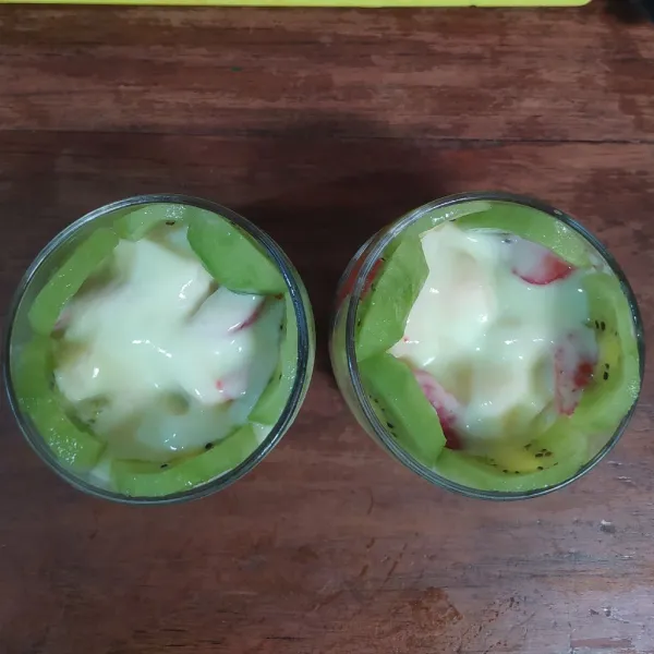 Tuang yogurt lalu tempel irisan kiwi pada dinding gelas, lalu beri kental manis.