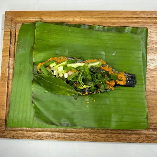 Tata ikan di atas daun pisang beri bumbu halus tambahkan tomat hijau iris, daun salam, sereh, daun bawang iris, kemangi.