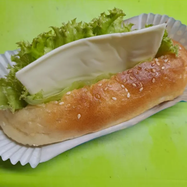 Belah memanjang roti hotdog bagian atasnya. Beri daun selada dan ½ bagian keju slice.