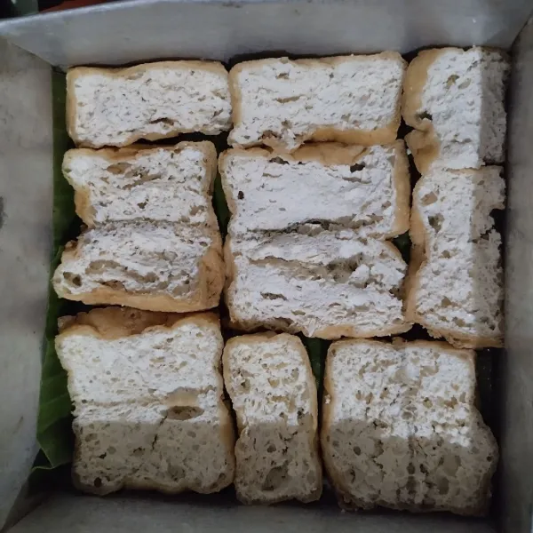 Tata tahu di dasar loyang yang sudah diolesi minyak dan dialasi daun pisang.