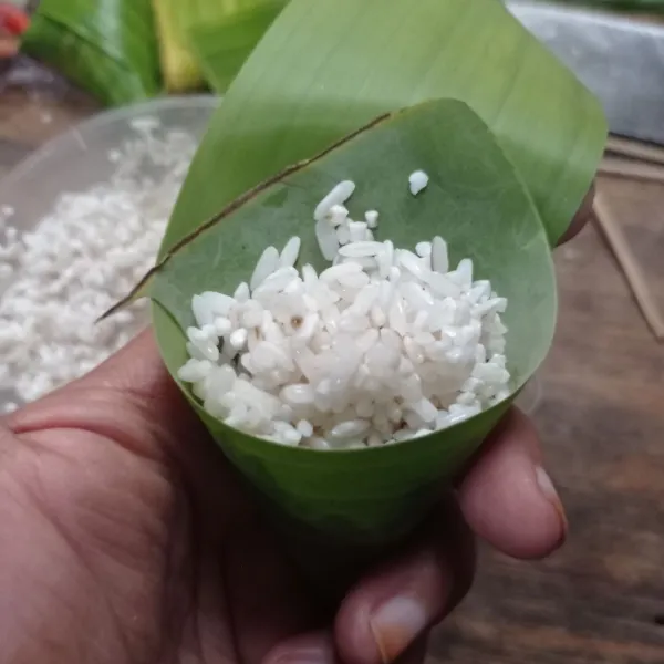 Siapkan daun pisang dengan ukuran 8 cm, lipat segitiga, kemudian isi dengan beras.