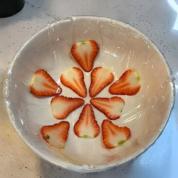 Lapisi bagian dalam mangkuk bulat sedang dengan plastik wrap. Iris strawberry tipis-tipis, lalu tata membentuk pola bunga di dasar mangkuk.