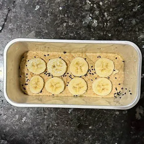 Oles tipis loyang dengan sedikit minyak, lalu tuang adonan ke dalamnya. Taburkan sedikit wijen hitam di atas adonan dan tata potongan pisang di permukaannya