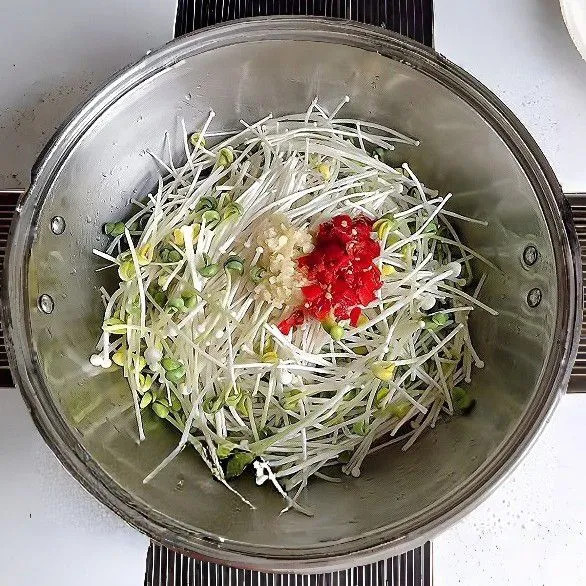 Masukkan kecambah kacang hitam dan jamur enoki ke dalam wajan. Tambahkan minyak sayur, lalu taburkan bawang putih cincang dan cabe merah cincang di atasnya.