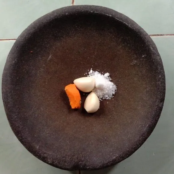 Haluskan kunyit, bawang putih, dan garam menggunakan ulekan.